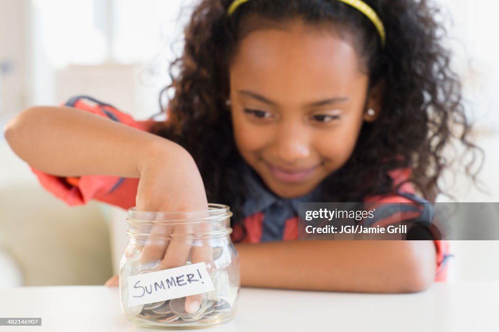 Mixed race girl putting coins in summer savings jar