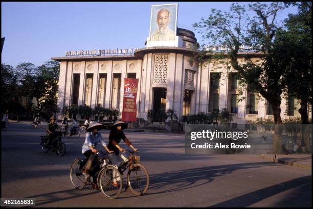 Cyclists ride past a poster of Ho Chi Minh set atop the opera house as the sun sets over Hanoi, Vietnam, 10th May 1983.