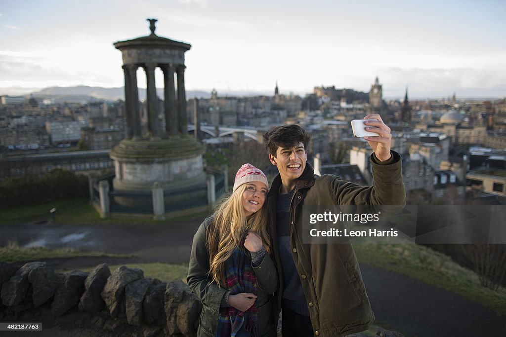 A young couple photograph themselves on Calton Hill with the background of the city of Edinburgh, capital of Scotland