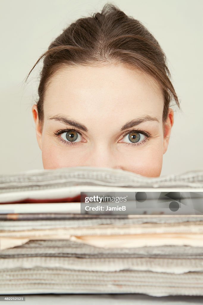 Head shot of young woman looking over the top of a stack of newspapers
