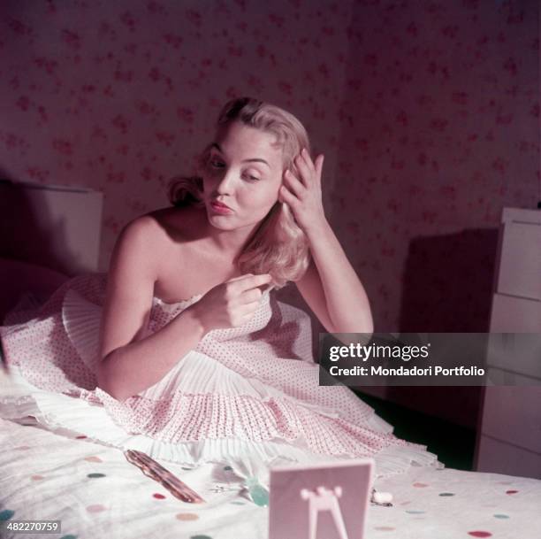 Miss Maria De Murtas in dot dress arranging her hair with a hairpin sitting on a bed. 1950s