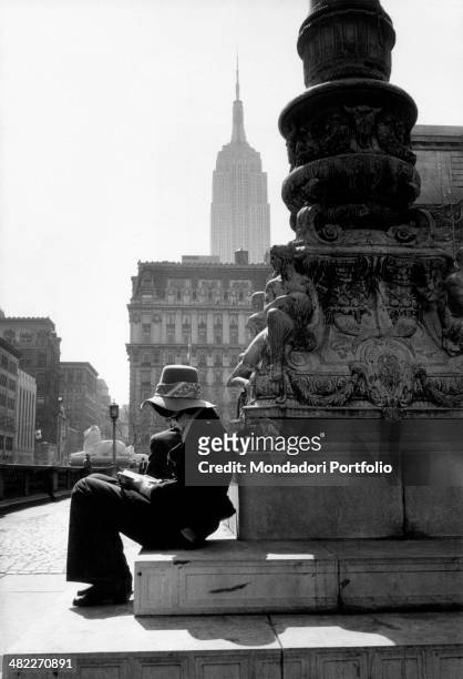 Student with a hat on her head reading sitting on the bronze base of the standard-bearer by Raffaele Menconi on the New York Public Library terrace....