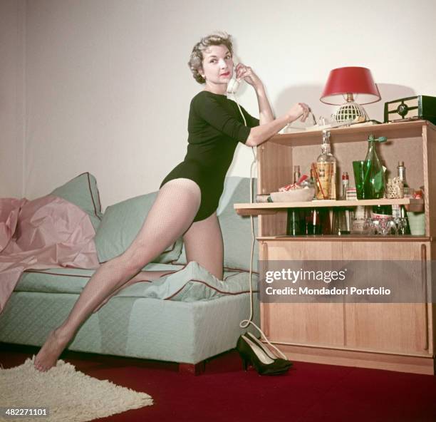 Woman in bodysuit and fishnet stockings posing leaning on a cocktail cabinet with a telephone in the hands. 1950s