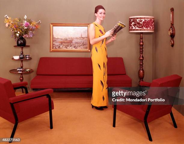 An Italian woman posing beside two armchairs and a design sofa wearing a yellow evening dress. Italy, 1960s
