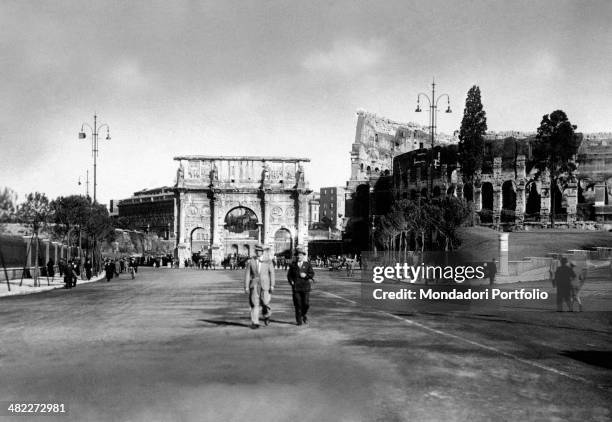 Two men walking along the via dell'Impero . Behind them, the Arch of Constantine and the Colosseum standing. Rome, 1930s