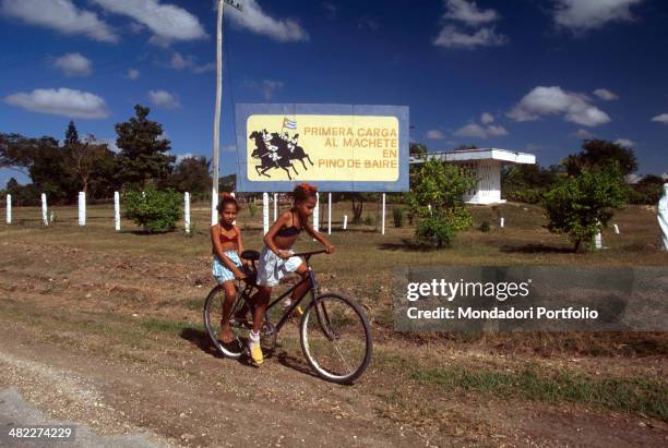 Two Cuban little girls riding bicycles. Behind them, the inscription on a poster Primera carga al machete en Pino de Baire remembering one of the...