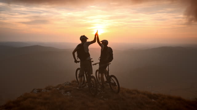 Aerial shot of two mountain bikers at the top of the mountain raising their hands and looking into the beautiful sunset. Shot in Slovenia, Europe.