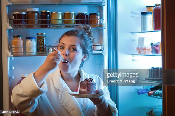 woman eating unhealthy chocolate cake in front of open refrigerator - 12 o'clock stock pictures, royalty-free photos & images