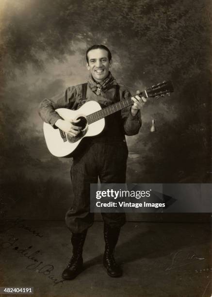 Portrait of an unidentified man, dressed in a overalls and a neckerchief as he smiles and plays an acoustic guitar, early 20th century. The picture...