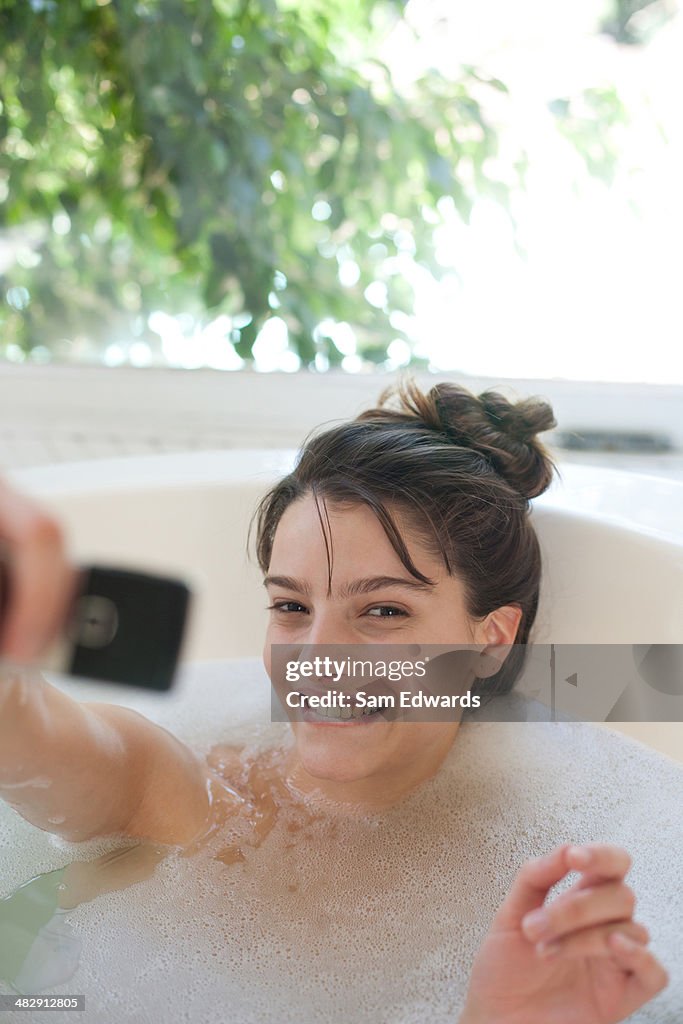 Woman relaxing in bubble bath using cellular phone and smiling