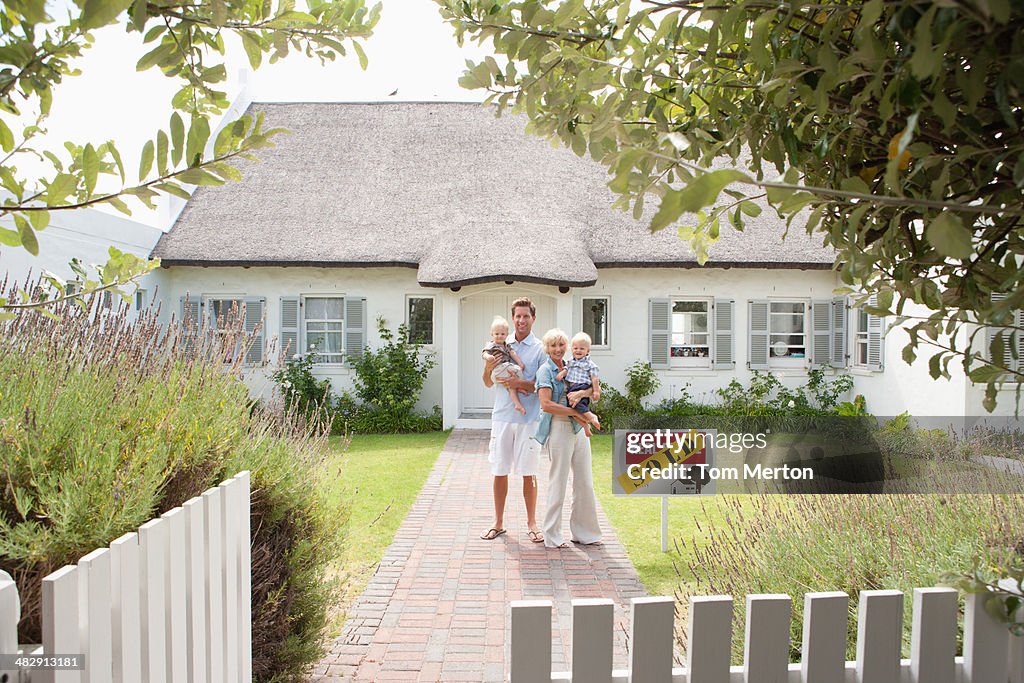 Man and woman holding babies in front of house with sold sign and white fence