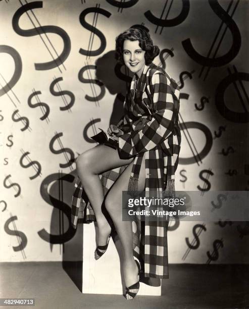 Portrait of American actress Frances Dee , in a checkered robe and with a pen in one hand, perched on a box in front of a wall that features dollar...
