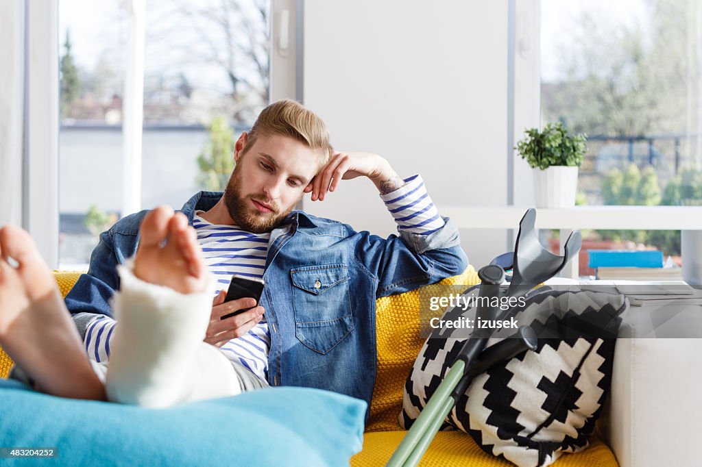 Junger Mann mit einem gebrochenen Bein mit Smartphone zu Hause fühlen