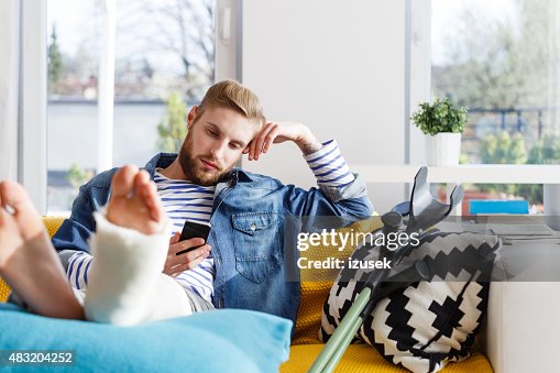 Junger Mann mit einem gebrochenen Bein mit Smartphone zu Hause fühlen Junger Mann mit einem gebrochenen Bein mit Smartphone zu Hause fühlen