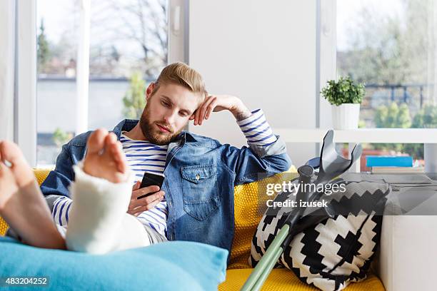 hombre joven con pierna fracturada usando teléfono inteligente en su casa - seguro médico fotografías e imágenes de stock