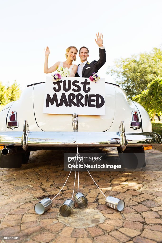 Newlyweds Waving In Convertible Car With Cans Attached To It