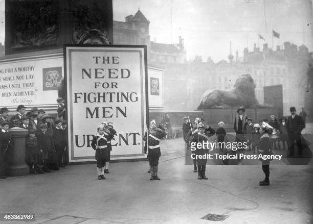 In Trafalgar Square, London street urchins dressed as soldiers with paper hats and canes as guns stand to attention watched by a small crowd. Behind...