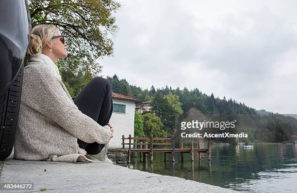 woman relaxes against car tire, lake edge - lake orta stock pictures, royalty-free photos & images