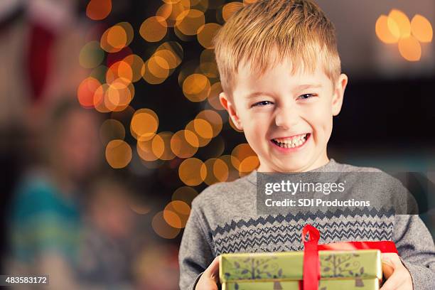 excited young boy holding christmas gift in front of tree - bois noel stock pictures, royalty-free photos & images