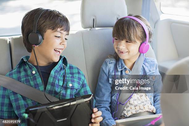 happy brother and sister with headphones using digital tablets in back seat of car - girls trip film stockfoto's en -beelden