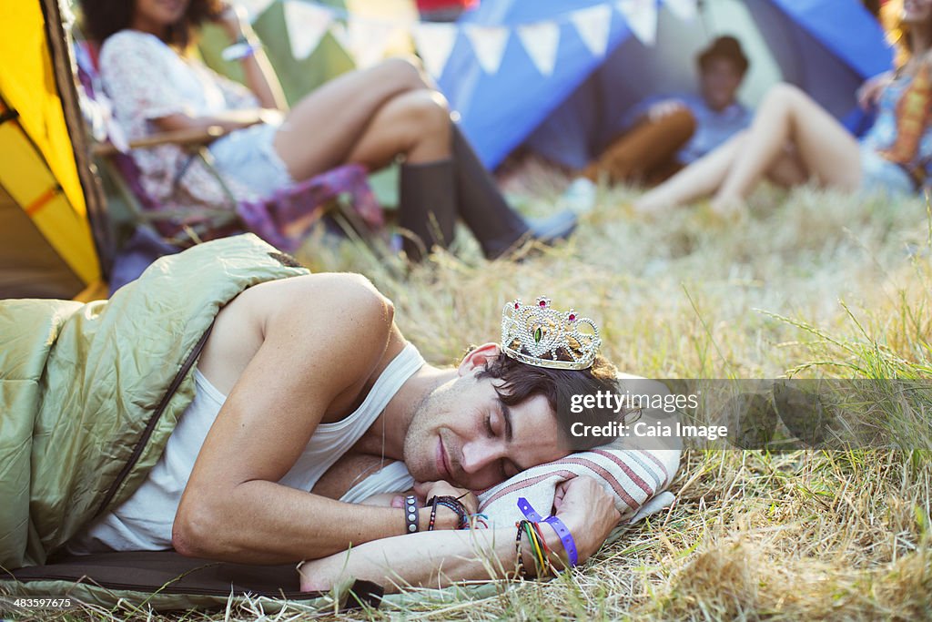 Man in tiara sleeping in sleeping bag outside tents at music festival