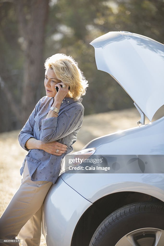 Woman talking on cell phone with automobile hood raised at roadside