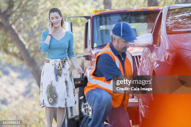 roadside mechanic fixing flat tire for woman on cell phone - car towing stock pictures, royalty-free photos & images