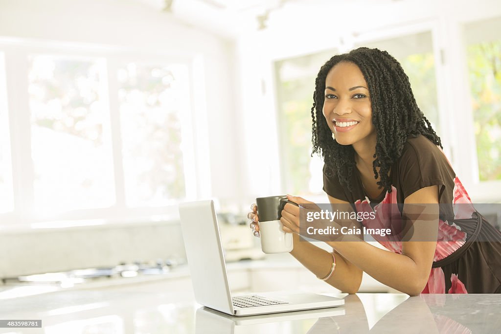 Portrait of smiling woman drinking coffee at laptop in kitchen