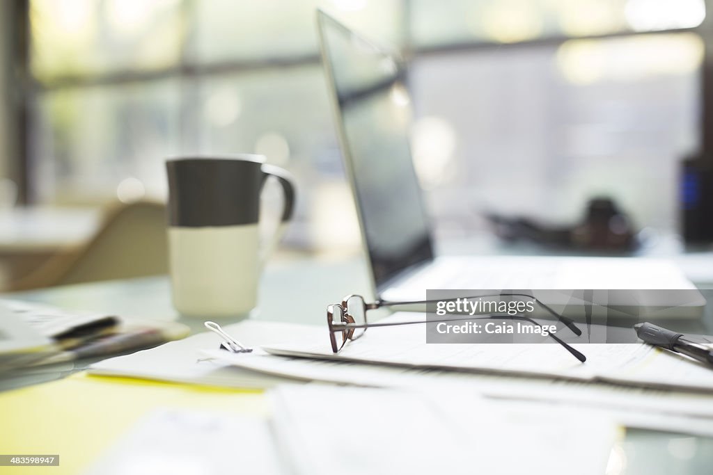Close up of coffee cup and eyeglasses on paperwork near laptop