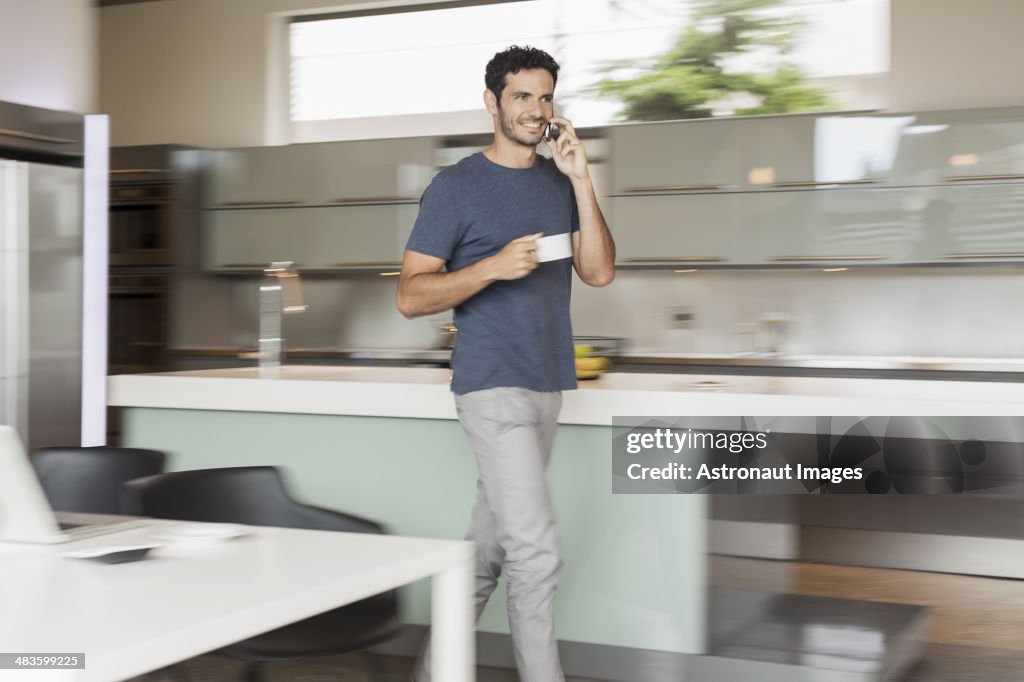 Smiling man drinking coffee and talking on cell phone in kitchen