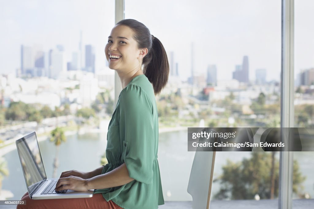 Portrait of confident casual businesswoman at laptop near window overlooking city