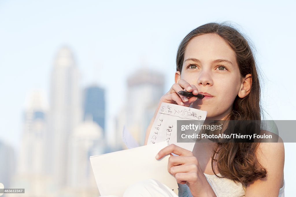Preteen girl sitting outdoors with pen in mouth, looking up in thought