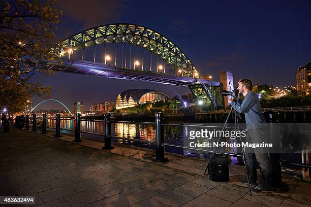 Portrait of a photographer taking pictures of the Tyne Bridge over the River Tyne at night, with the Gateshead Millennium Bridge visible in the...