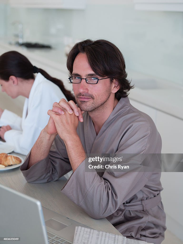 Man in robe with eyeglasses sitting with laptop and woman in background