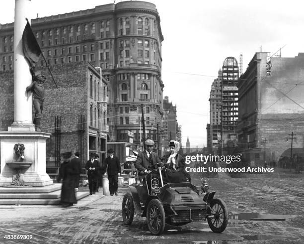 Couple in their car on Market Street with the Ferry Building in the background, San Francisco, California, early to mid 1900s.