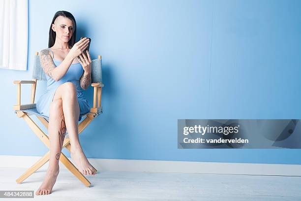 tattooed girl wearing a blue dress looking at her phone - directors chair stock pictures, royalty-free photos & images