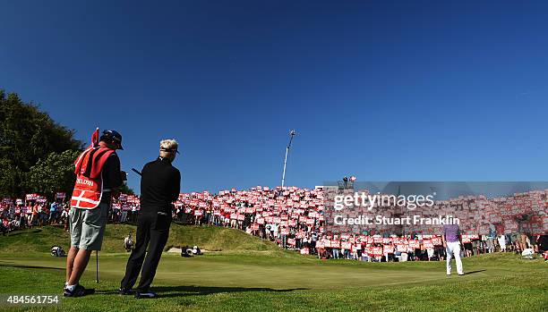 Soren Kjeldsen of Denmark watches on the 16th green as fans hold placards to celebrate the 500th tournement during the first round of the Made in...