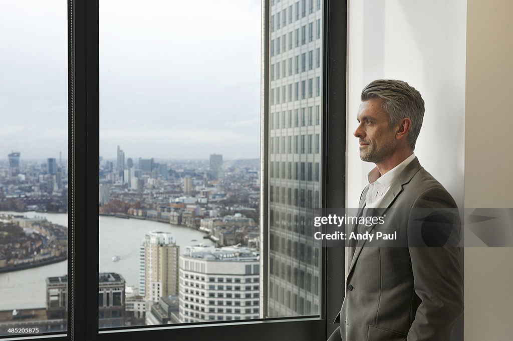 Portrait of mature businessman looking out of office window, Canary Wharf, London, UK