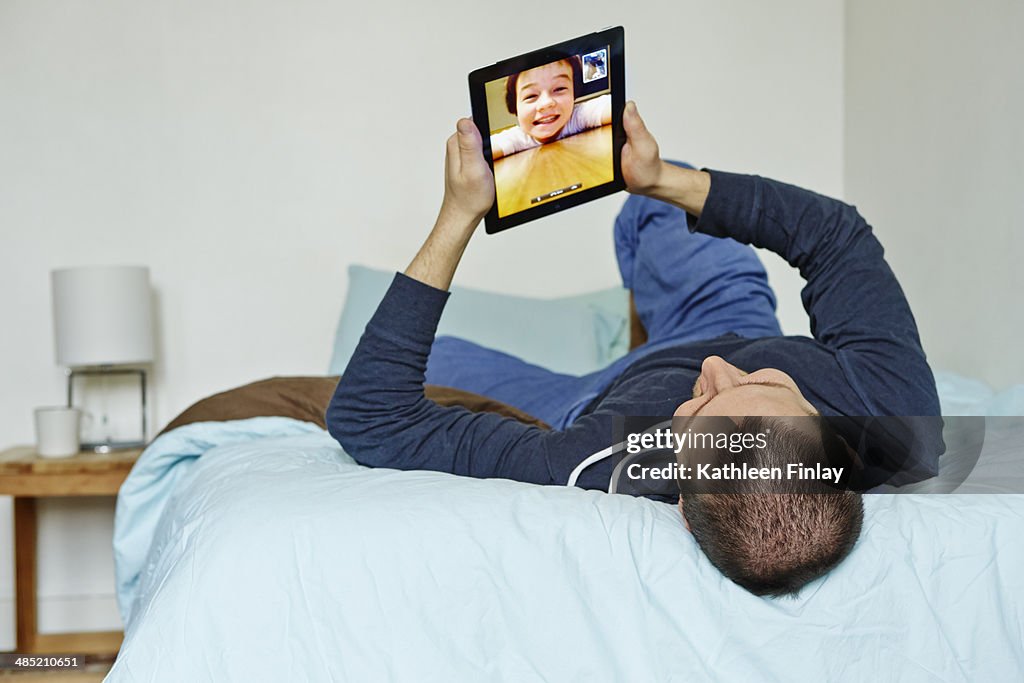 Mid adult man lying on bed holding up digital tablet