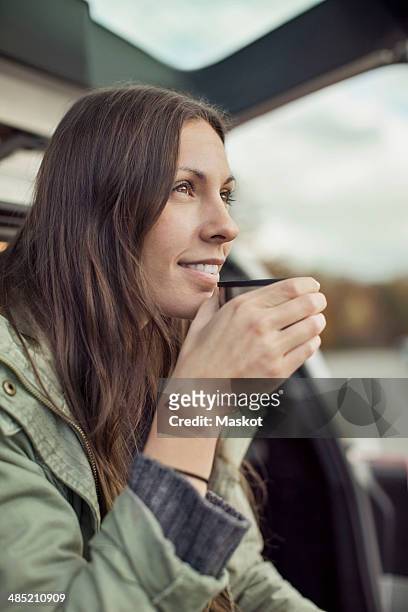 young woman looking away while drinking coffee at car's trunk - cafeïne stockfoto's en -beelden