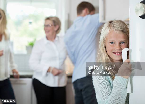 portrait of smiling girl holding door handle with family in background at kitchen - fridge handle stock pictures, royalty-free photos & images