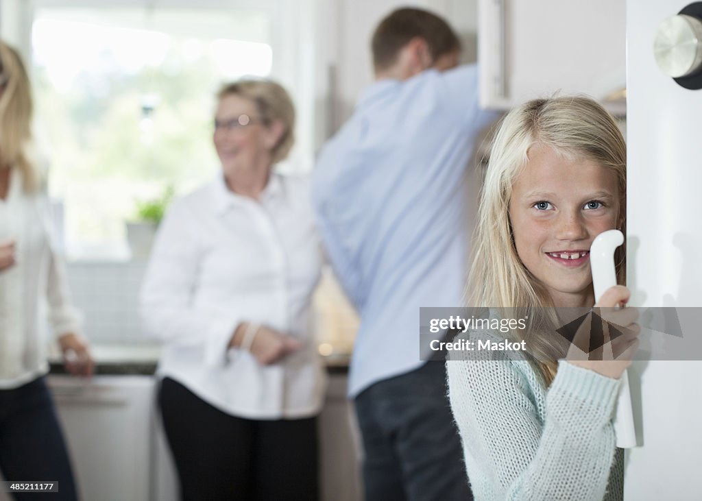 Portrait of smiling girl holding door handle with family in background at kitchen