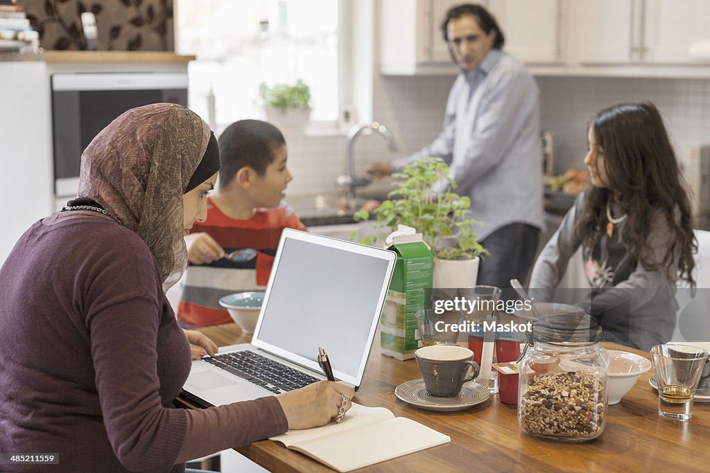 Muslim woman working on laptop with family having breakfast in kitchen