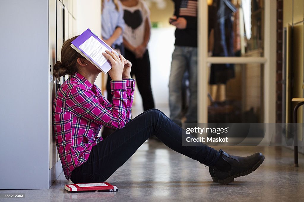Side view of bored schoolgirl covering face with book in locker room