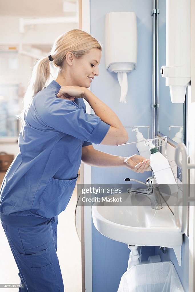 Side view of young nurse using soap dispenser to wash hands in bathroom