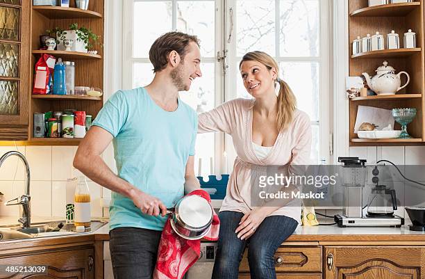 man wiping saucepan while looking at woman sitting on kitchen counter - sober leven stockfoto's en -beelden