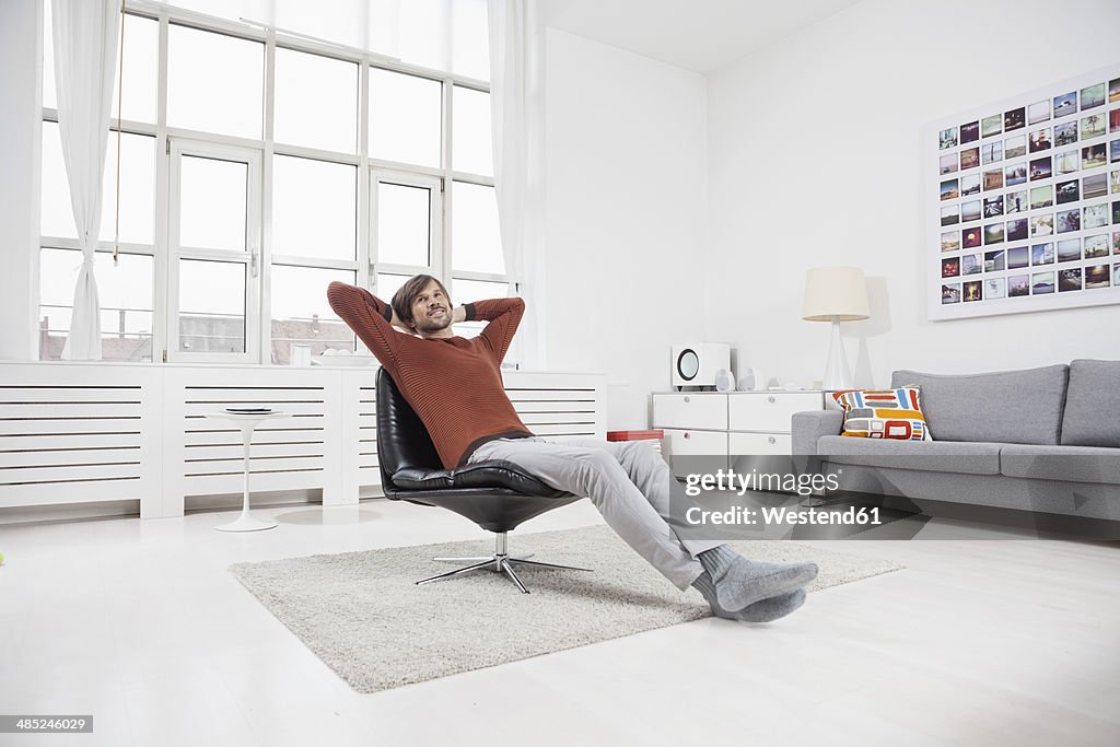 Germany, Munich, Man at home, sitting in chair, hands behind head