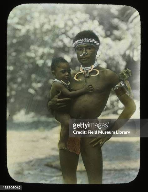 Portrait of a man in tapa loin cloth and boar's tusk necklace, holding a young boy in his arms, Ragetta Island, Papua New Guinea, January 1910.