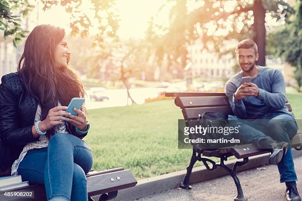 flirting couple in the park texting on smartphones - lust girl stock pictures, royalty-free photos & images