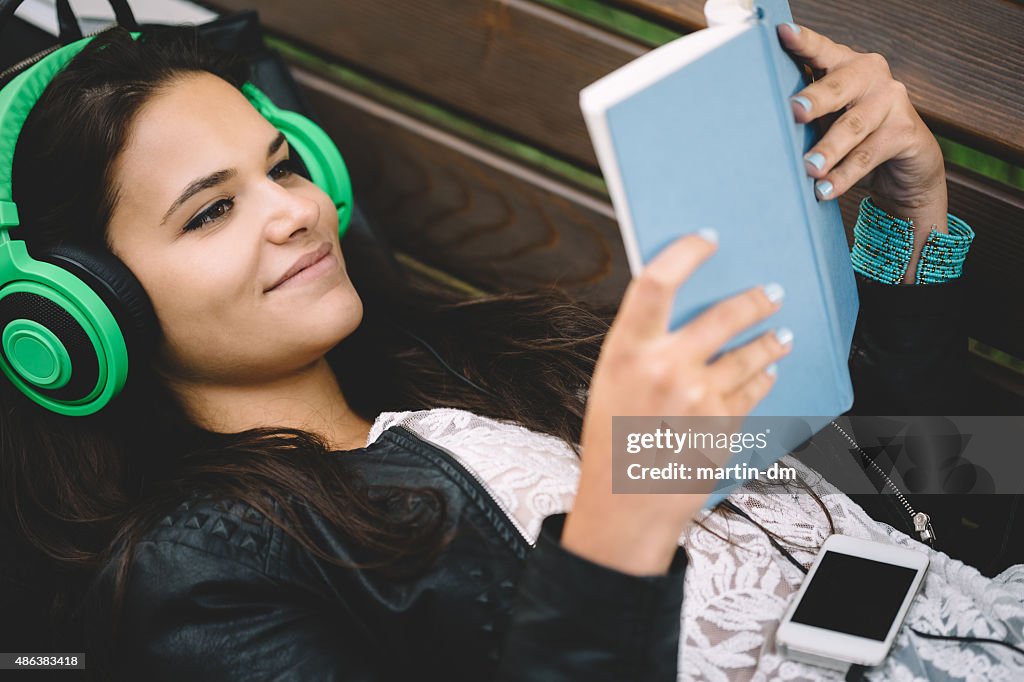 Schoolgirl with headphones and book resting on bench